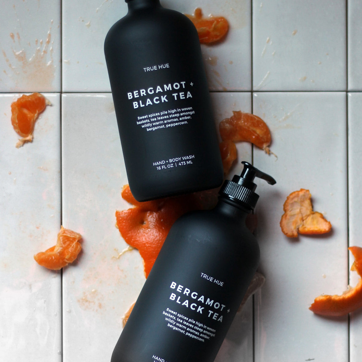 two bottles of bergamot + black tea hand soaps over a white tiled background with mandarin oranges.