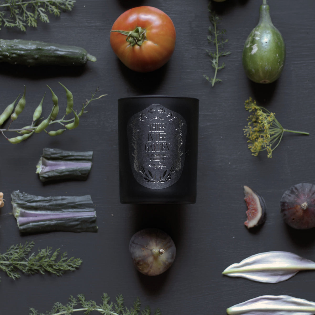 Thief In The Garden Candle on black background with vegetables, flower petals and leaves next to the candle.