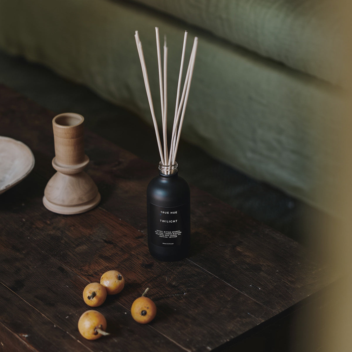 Twilight Reed Diffuser sitting on a wood coffee table next to a candle stick holder and a few small, orange fruits.