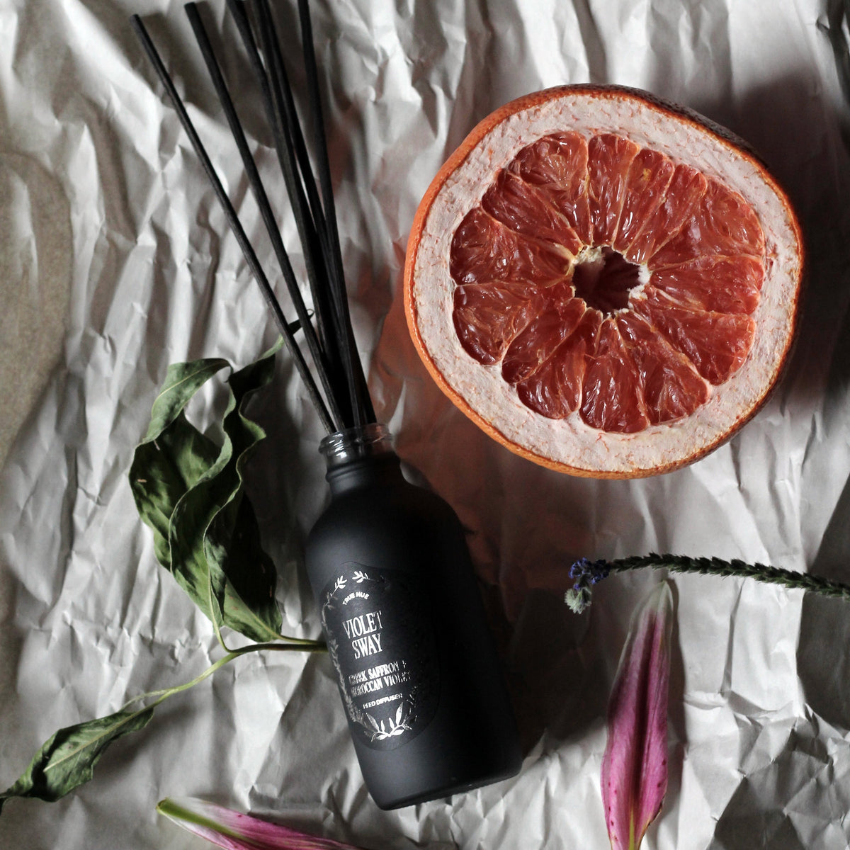 Violet Sway Reed Diffuser laying on white paper background next to flower petals, leaves and a cut piece of grapefruit.