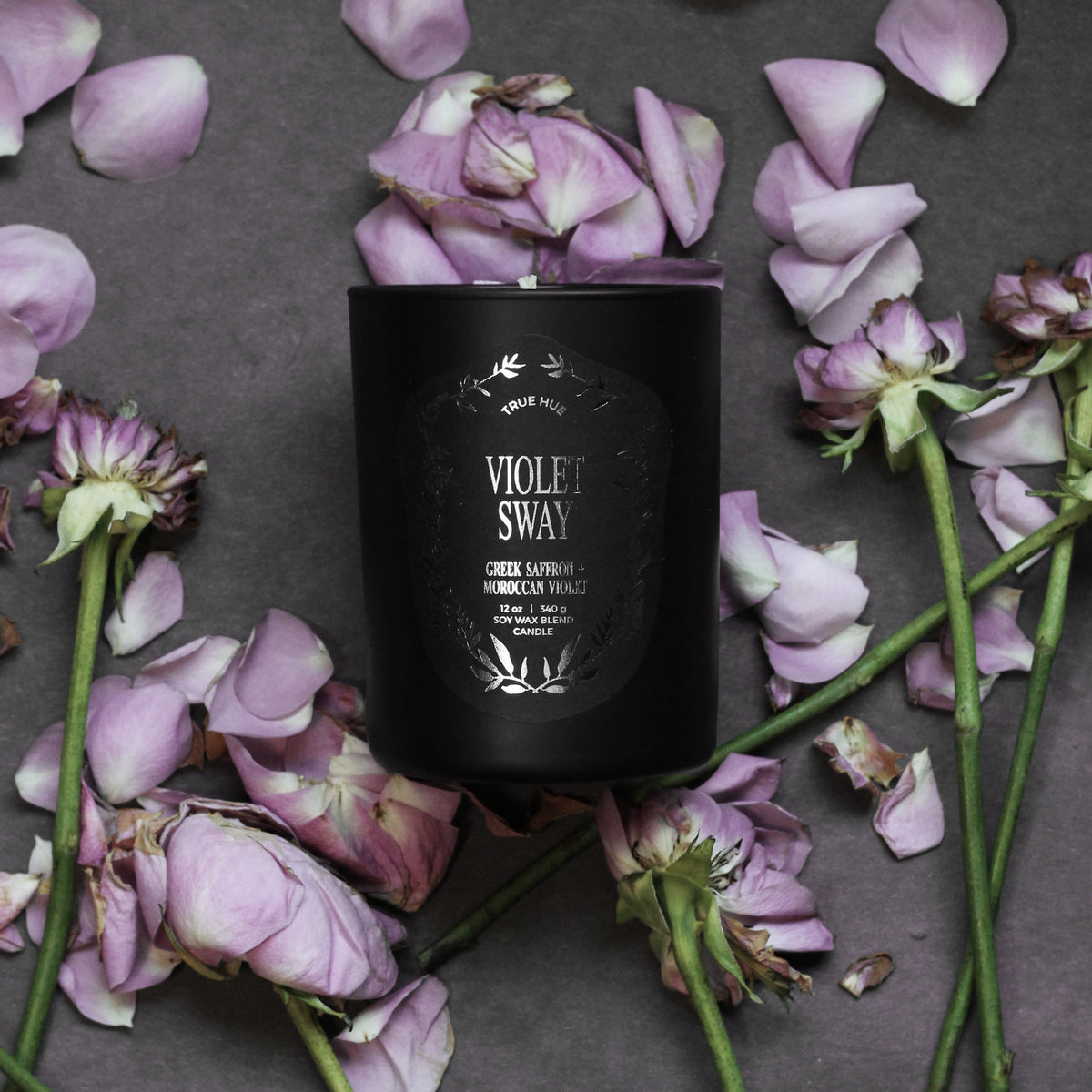 Black candle labeled 'Violet Sway' surrounded by purple flowers on a dark surface