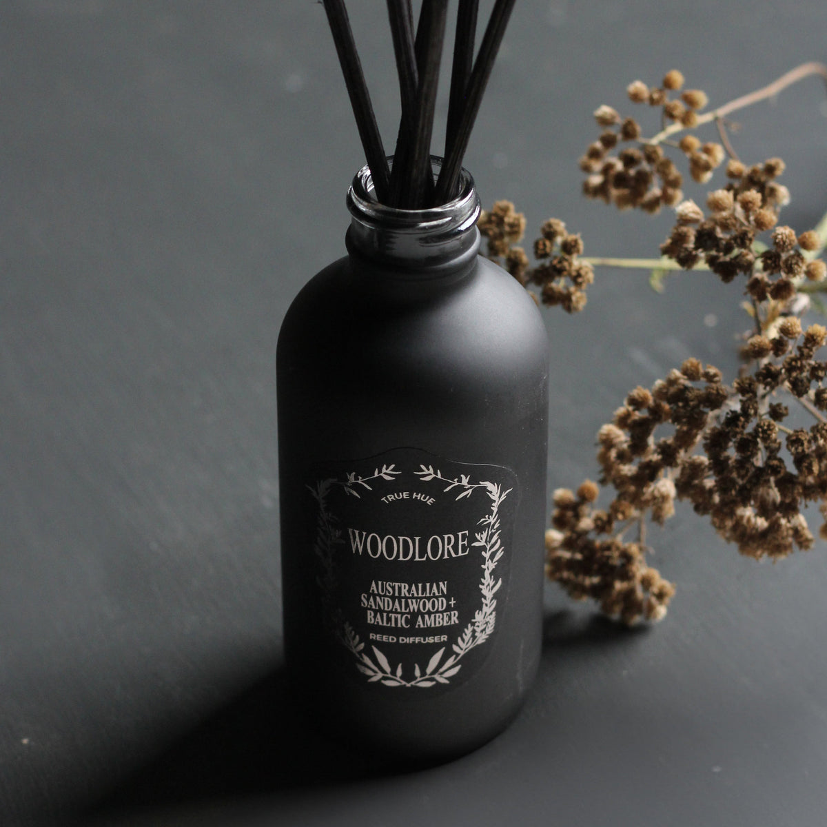 Woodlore Reed Diffuser close up on a black background with floral sprig next to the bottle.