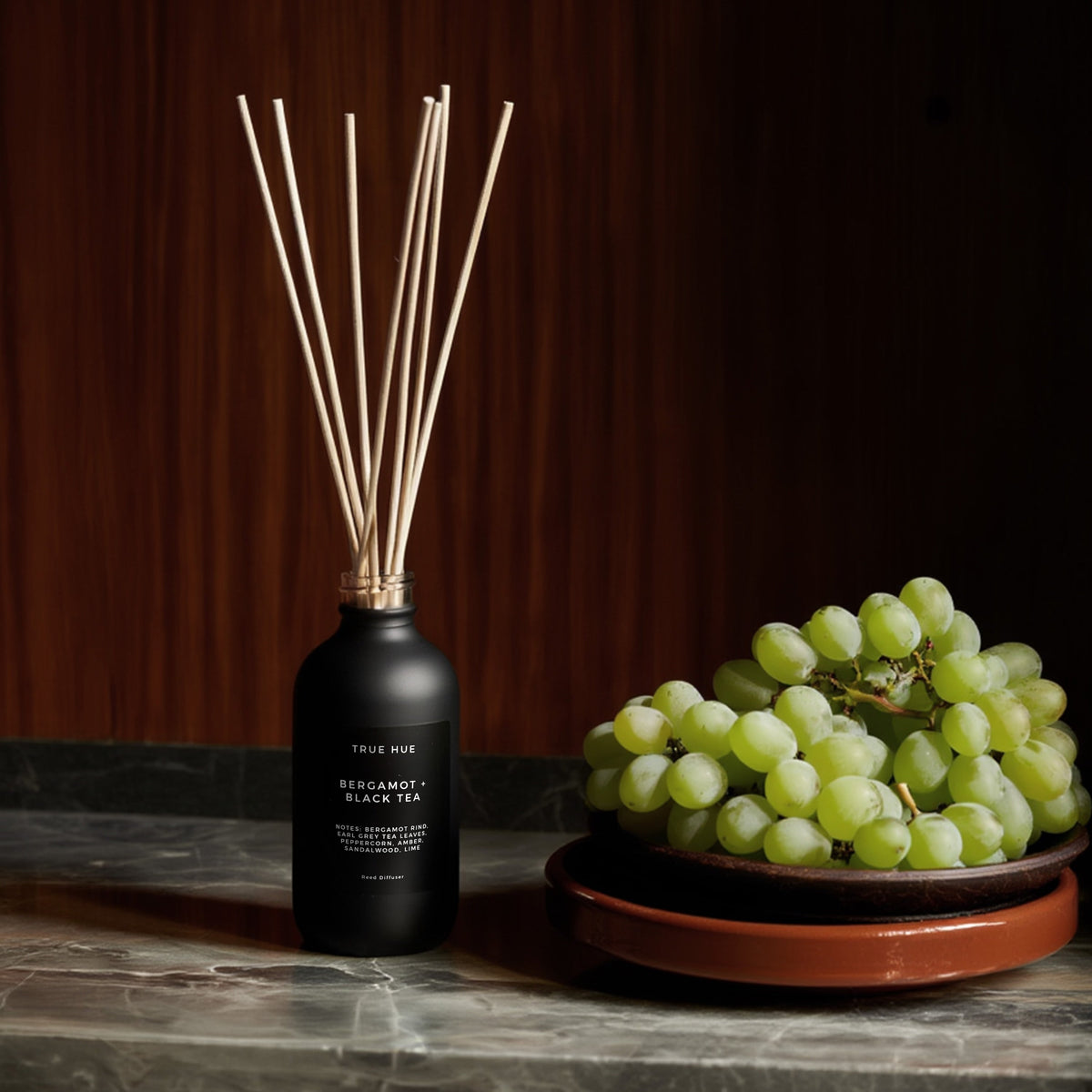 Bergamot + Black Tea Reed Diffuser sitting on a marble countertop next to a bowl of green grapes.
