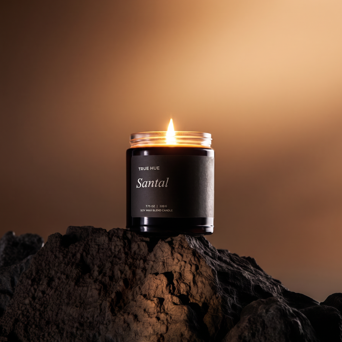 A lit candle in a black glass jar with a black label reading 'TRUE MUE Santal' sits on dark textured rocks against a warm brown gradient background.