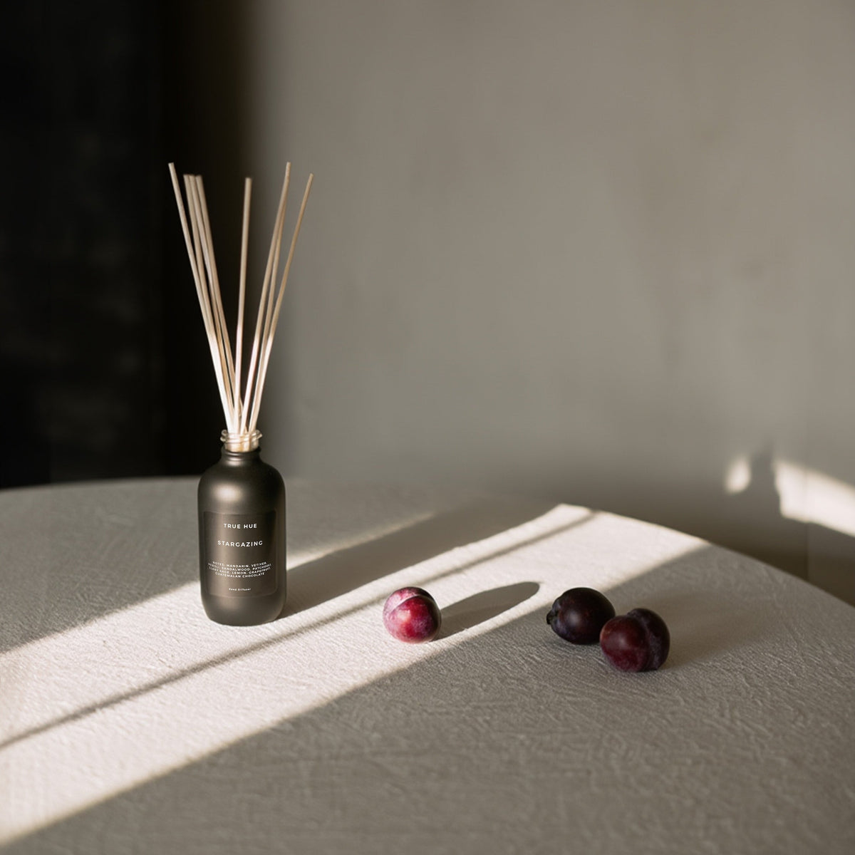 Stargazing Reed Diffuser sitting on a round table with a beige table cloth and three plums sitting next to the diffuser.
