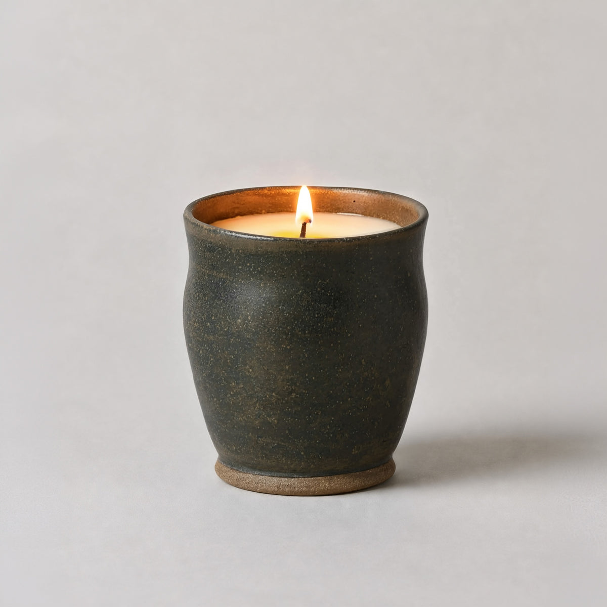 A lit candle in a dark, textured ceramic pot with a warm brown interior and base, set against a plain light gray background.