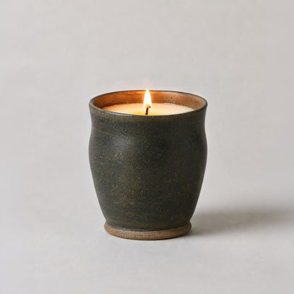 A lit candle in a dark, textured ceramic pot with a warm brown interior and base, set against a plain light gray background.