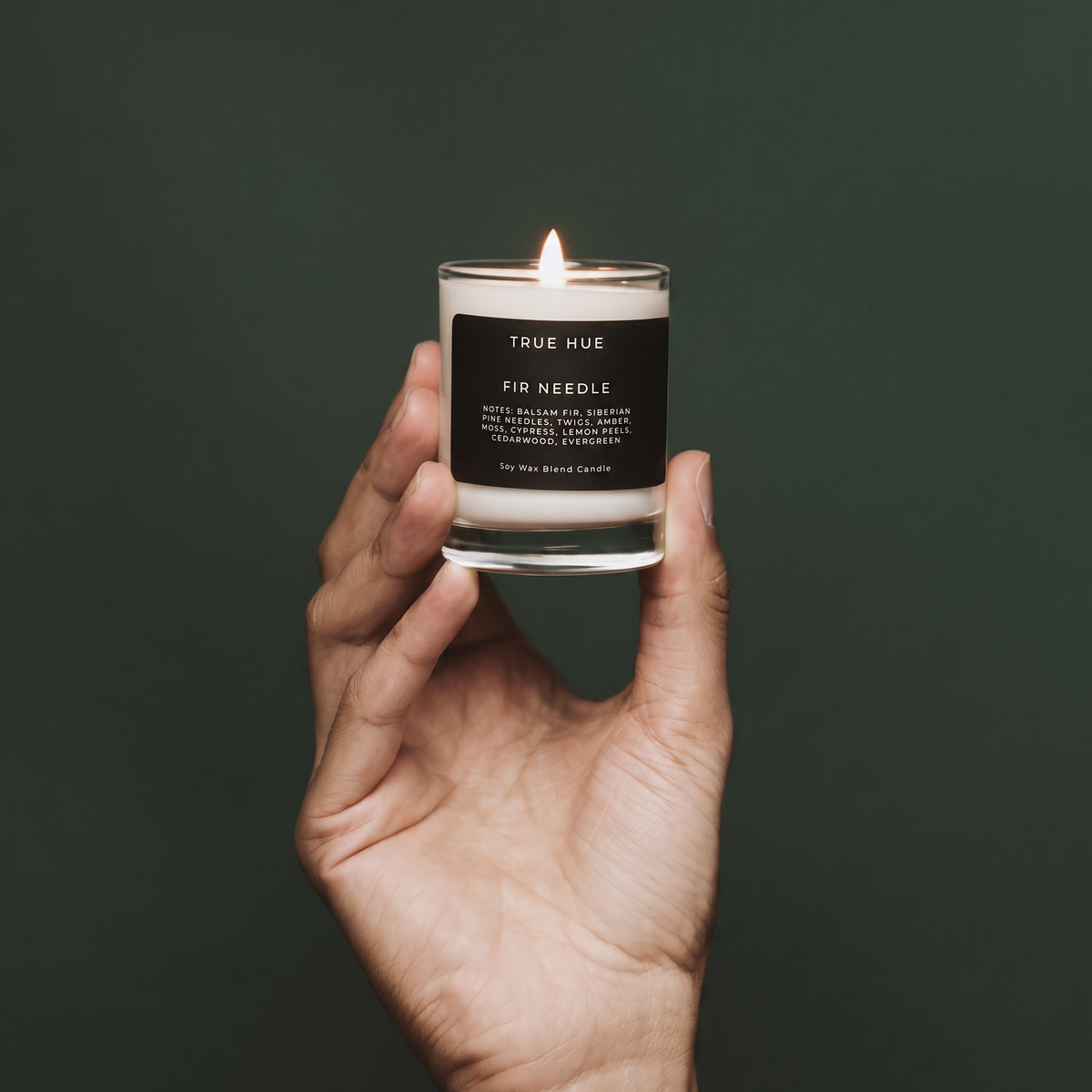 A hand holding a lit glass jar candle against a dark green background. The candle label reads 'TRUE HUE' and 'PIN NEEDLE'.