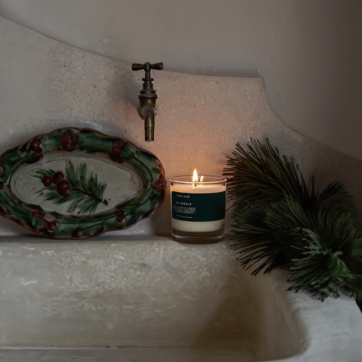 A lit candle in a clear glass jar sits on the edge of a stone sink, surrounded by a decorative holiday plate, a brass faucet, and green pine branches.